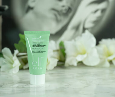 A travel size tube of the e.l.f. Blemish Breakthrough Blemish Calming Water Cream, green with a white lid, standing in front of a dark background with white flowers