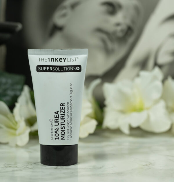 A black and white tube of The Inkey List 10% Urea Moisturizer - Rough and Dry Skin Solution standing in front of a dark background with white flowers