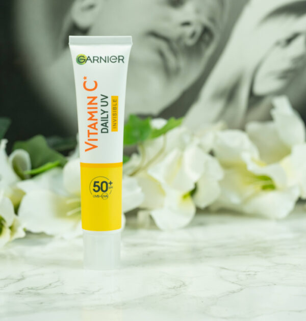A tube of Garnier Vitamin C Daily UV Invisible SPF 50+ standing in front of a dark background with white flowers