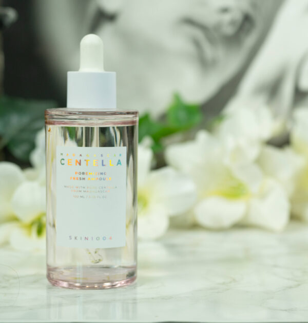 A light pink bottle of SKIN1004 Madagascar Centella Poremizing Fresh Ampoule standing in front of a dark background with white flowers