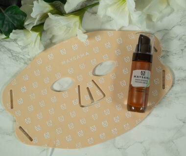 The Maysama Prana Pulsed LED Light Therapy Mask with a bottle of Green Rooibos Pressed Serum lying on a marbled surface. The mask is light brown and has several small Maysama logos in white on it.