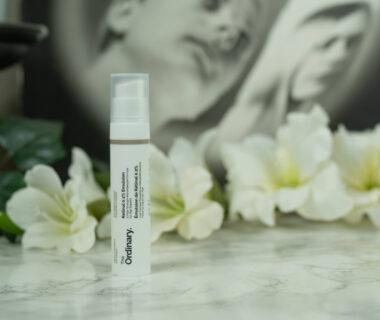 A tube of The Ordinary Retinal 0.2% Emulsion standing in front of a dark background with white flowers