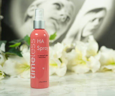 A bottle of Timeless HA Rose Spray with Matrixyl 3000 standing in front of a dark background with white flowers