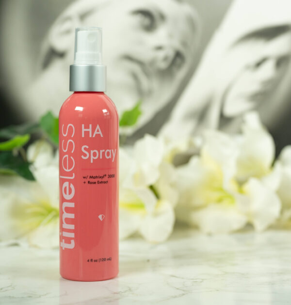 A bottle of Timeless HA Rose Spray with Matrixyl 3000 standing in front of a dark background with white flowers