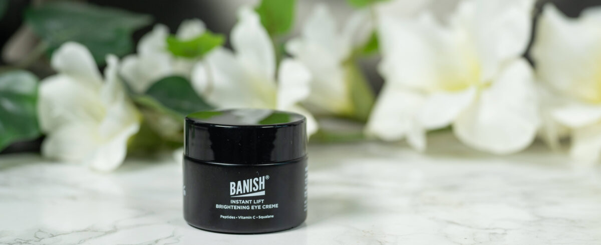 A black pot of Banish Instant Lift Brightening Eye Cream standing in front of a dark background with white flowers. It has white writing.