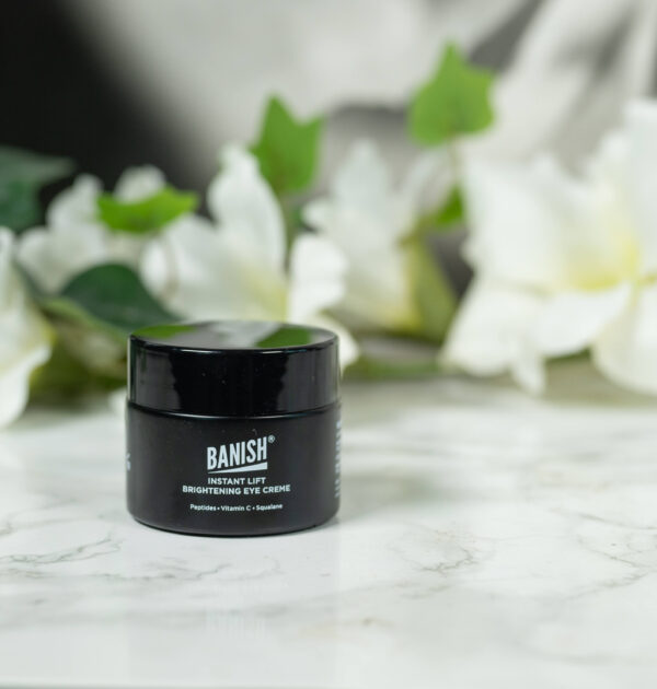 A black pot of Banish Instant Lift Brightening Eye Cream standing in front of a dark background with white flowers. It has white writing.