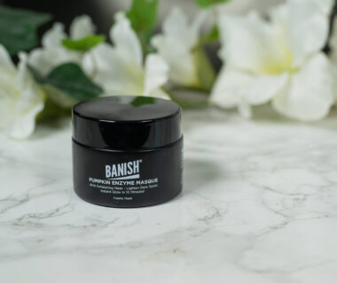 A pot of Banish Pumpkin Enzyme Mask standing in front of a dark background with white flowers