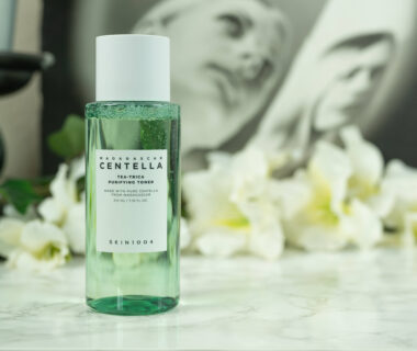 A bottle of SKIN1004 Tea-Trica Purifying Toner standing in front of a dark background with white flowers