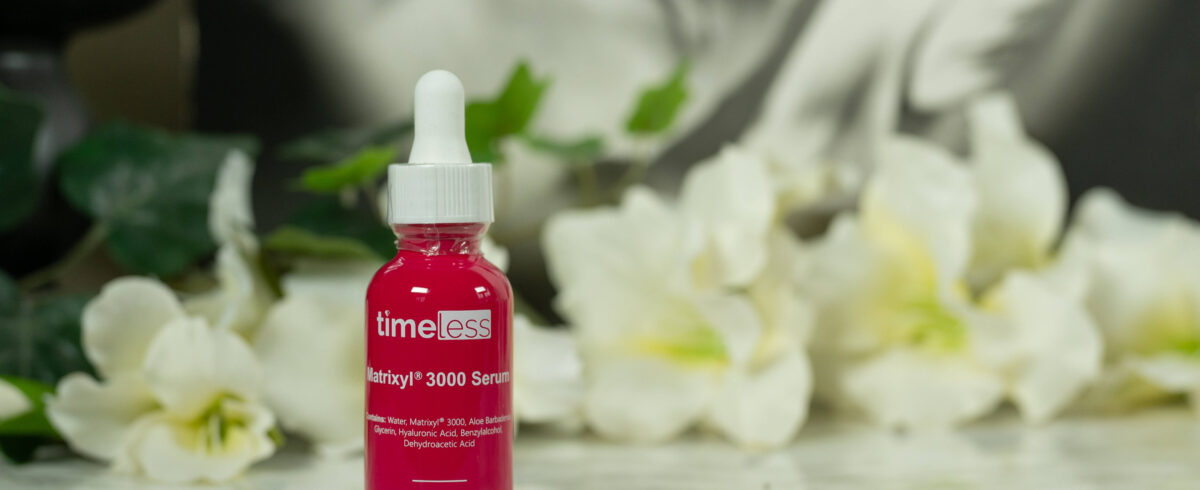 A bottle of timeless Matrixyl 3000 Serum in a deep rose color with white writing, standing in front of a dark background with white flowers