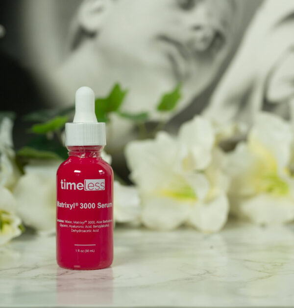 A bottle of timeless Matrixyl 3000 Serum in a deep rose color with white writing, standing in front of a dark background with white flowers