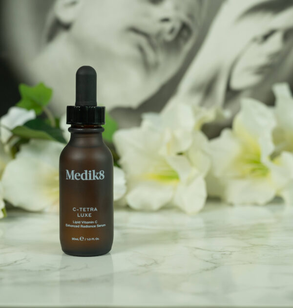 A bottle of Medik8 C-Tetra Luxe Serum, made from brown frosted glass, tandin gin front of a dark background with white flowers