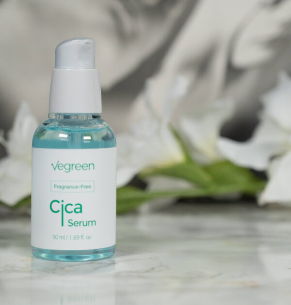 A bottle of Vegreen Fragrance-Free Cica Serum (old packaging) standing in front of a dark background with white flowers